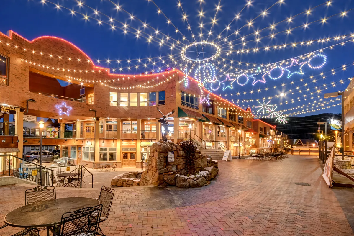 An outdoor scene featuring decorative lights, a stone fountain, outdoor seating, and inviting shop facades, with a festive, joyful ambiance. People appear to be gathering and enjoying the outdoor environment, suggesting a lively community with events and al fresco dining.