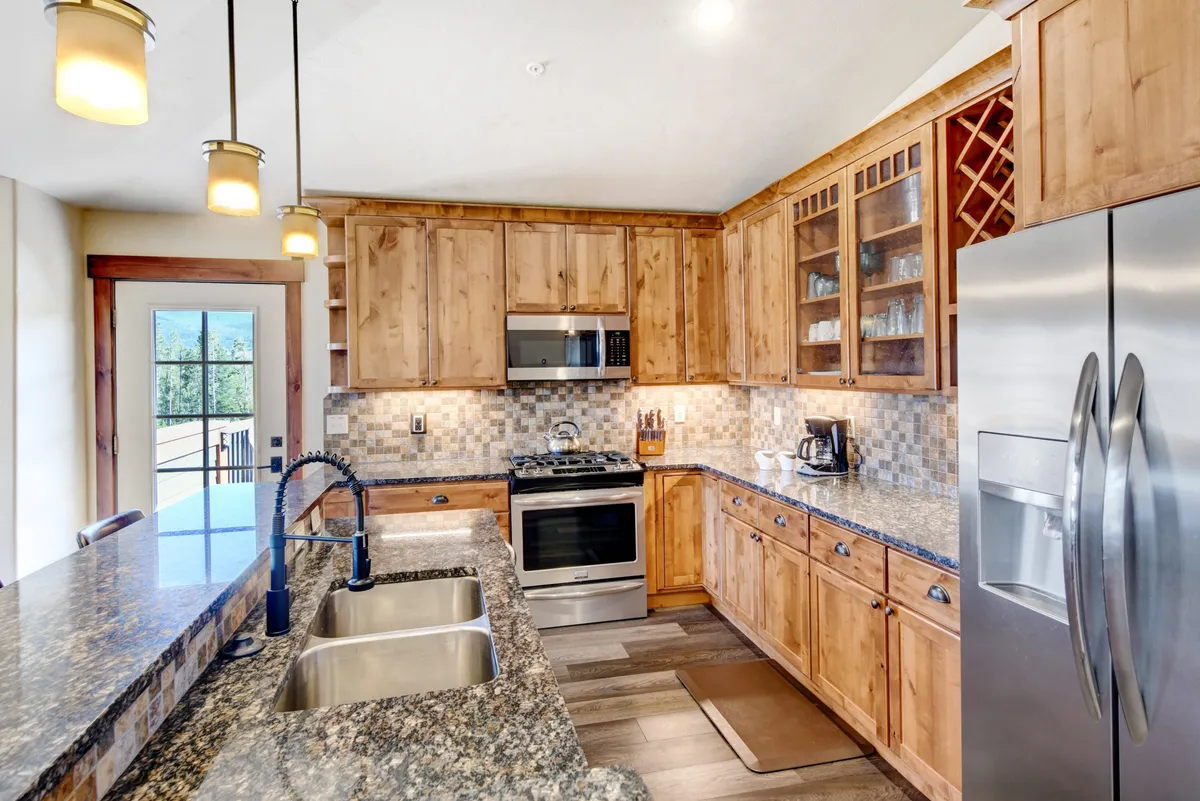 A warmly lit gourmet kitchen featuring expansive granite countertops, elegant wood cabinetry, and high-end stainless steel appliances. The kitchen island with a built-in sink offers generous workspace and casual bar seating, ideal for both preparing meals and socializing. The layout is open and welcoming, designed for both culinary inspiration and family gatherings.