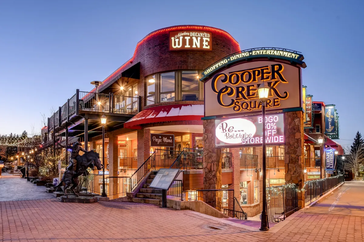 The image showcases the lively entrance to Cooper Creek Square with festive lighting, a wine shop sign, and broad stairs leading into a bustling local destination. Decorative touches and evening ambiance suggest a vibrant, family-friendly hub for shopping, dining, and entertainment.