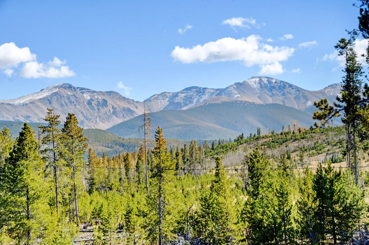 An expansive exterior view of the Rocky Mountains, framed by lush green trees and a blue sky with scattered clouds. The scene evokes possibilities for hiking, biking, and outdoor exploration during spring, summer, and fall.