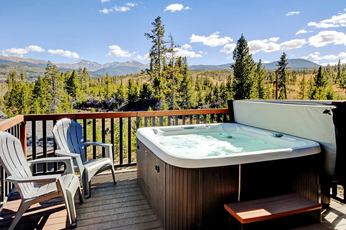 This exterior image captures a private hot tub set on a scenic patio, flanked by adirondack chairs and oriented towards sweeping mountain views. The tranquil outdoor setting is bathed in gentle natural light, suggesting a perfect spot for relaxation or romantic evenings under the stars.