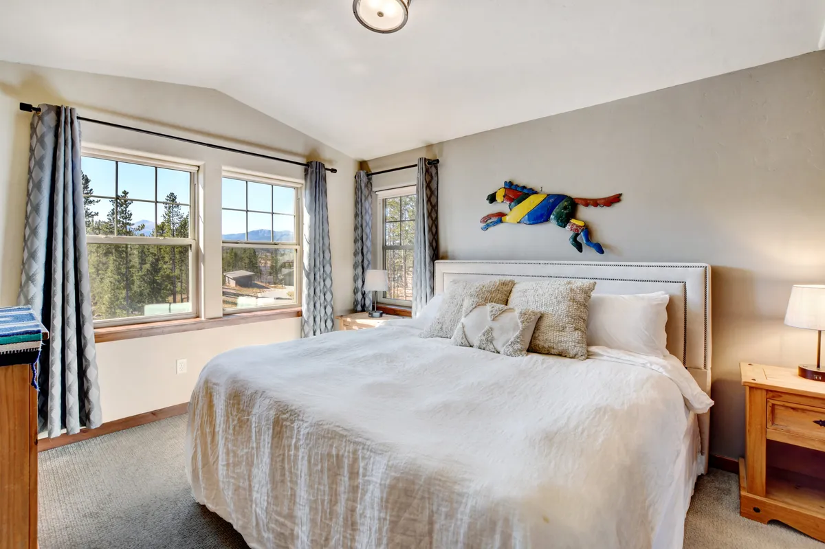 A bright bedroom with white linen bedding, colorful wall decoration, and large windows flooding the space with natural light. The room feels inviting, tranquil, and perfect for relaxing or starting the day with scenic views.