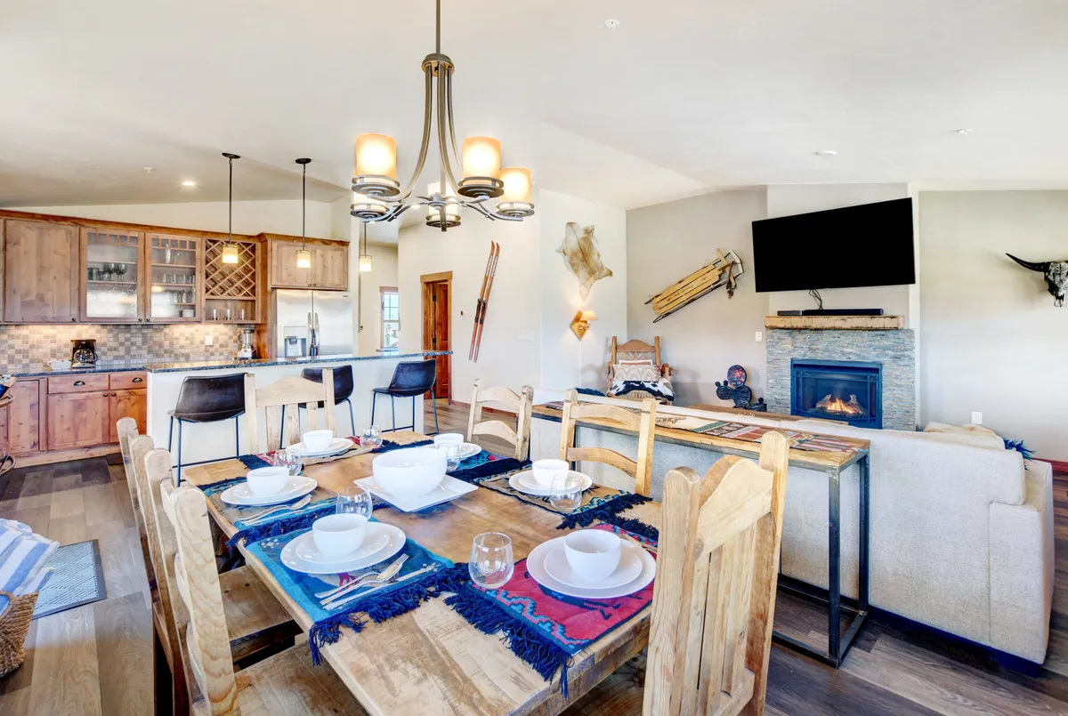 A cozy dining area set with decorative place settings, adjacent to a modern fireplace and kitchen. The ambiance is welcoming and perfect for shared meals or entertaining guests after a day outdoors.